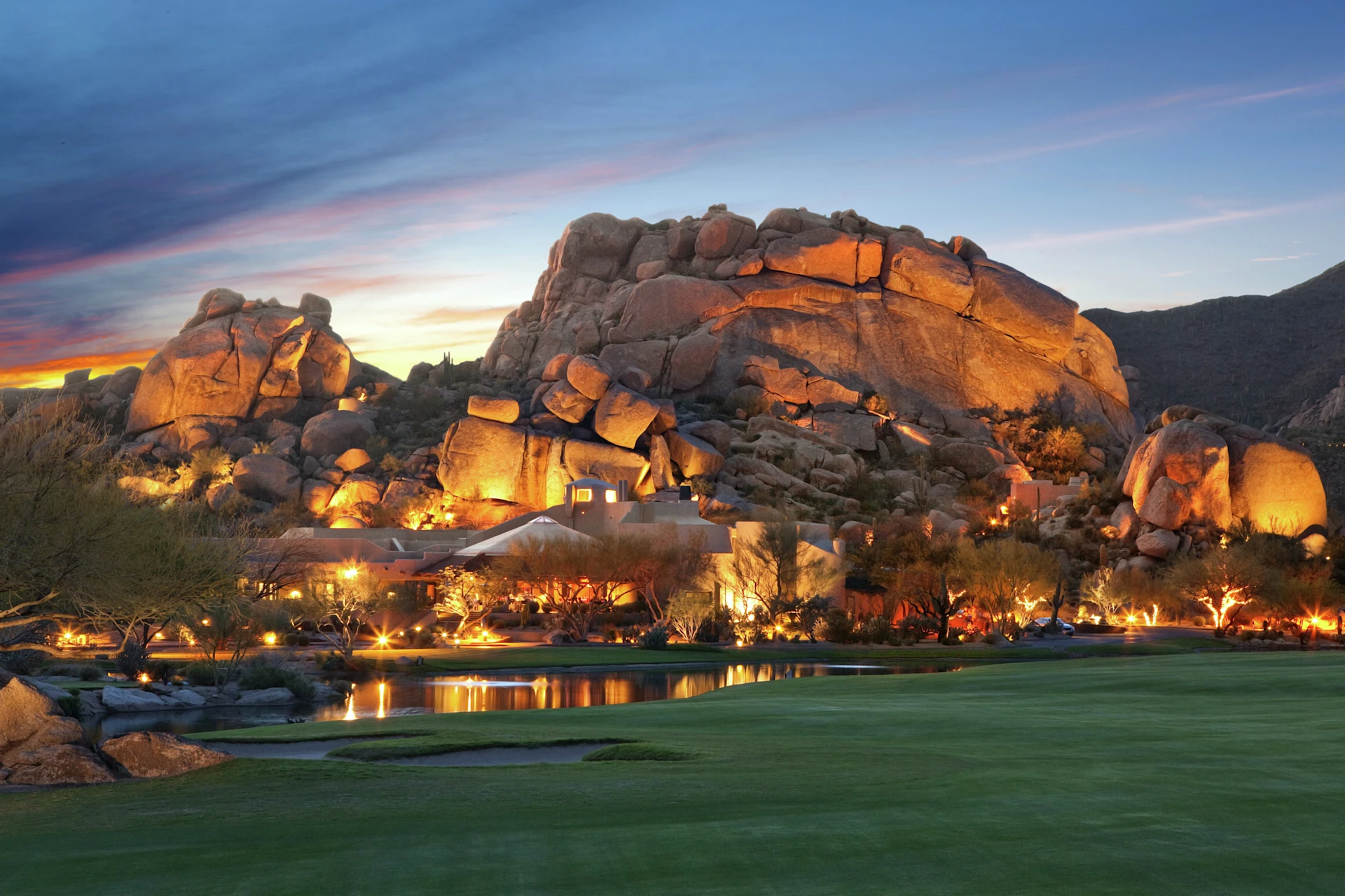 Desert resort at sunset with illuminated rock formations, trees, a pond, and a manicured green lawn in the foreground.