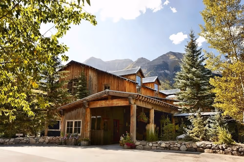 Rustic wooden lodge surrounded by evergreen and deciduous trees with mountains in the background under a sunny sky.