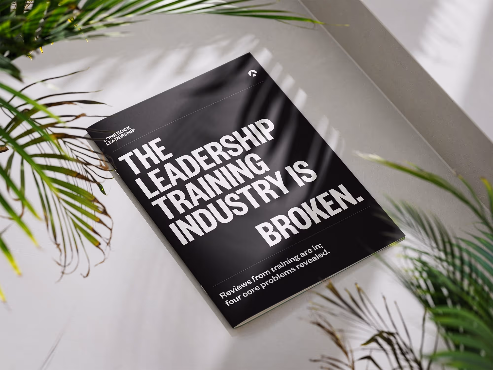 Black booklet titled 'The Leadership Training Industry is Broken' lying on a white surface surrounded by green plant leaves.