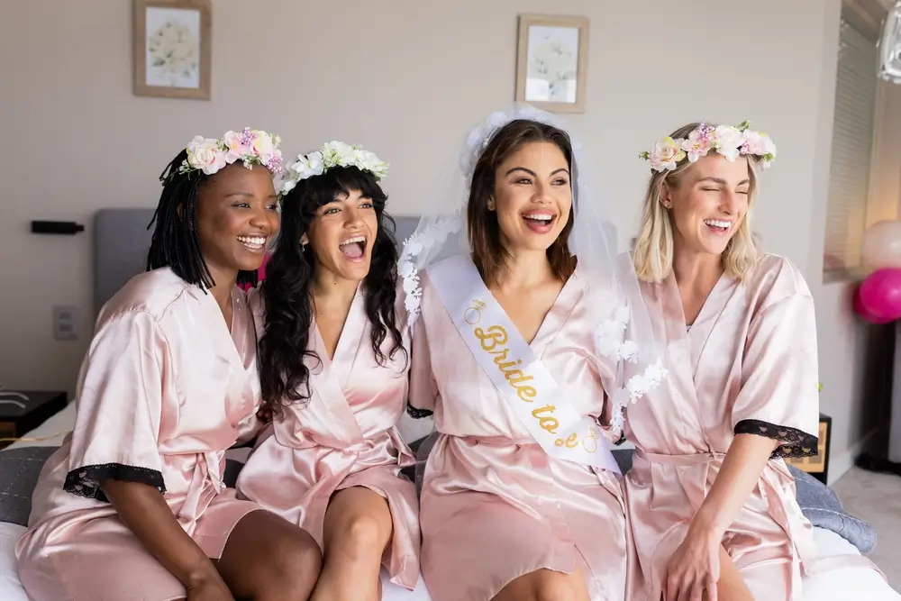 Bride-to-be with friends wearing robes and flower crowns, laughing on bed