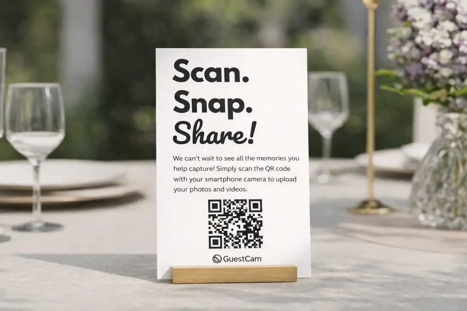 A GuestCam 'Scan. Snap. Share.' QR code sign displayed in a wooden table holder, surrounded by wine glasses and floral centerpieces.