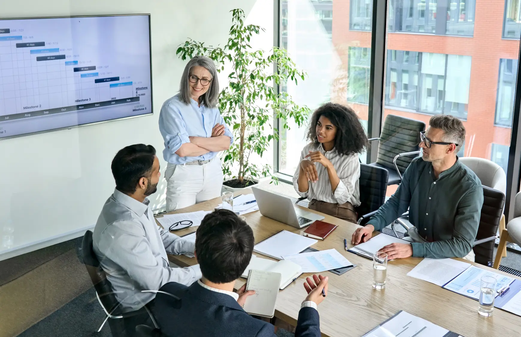 A marketing or communications team meeting in a modern conference room, with a project timeline displayed on a screen behind them.