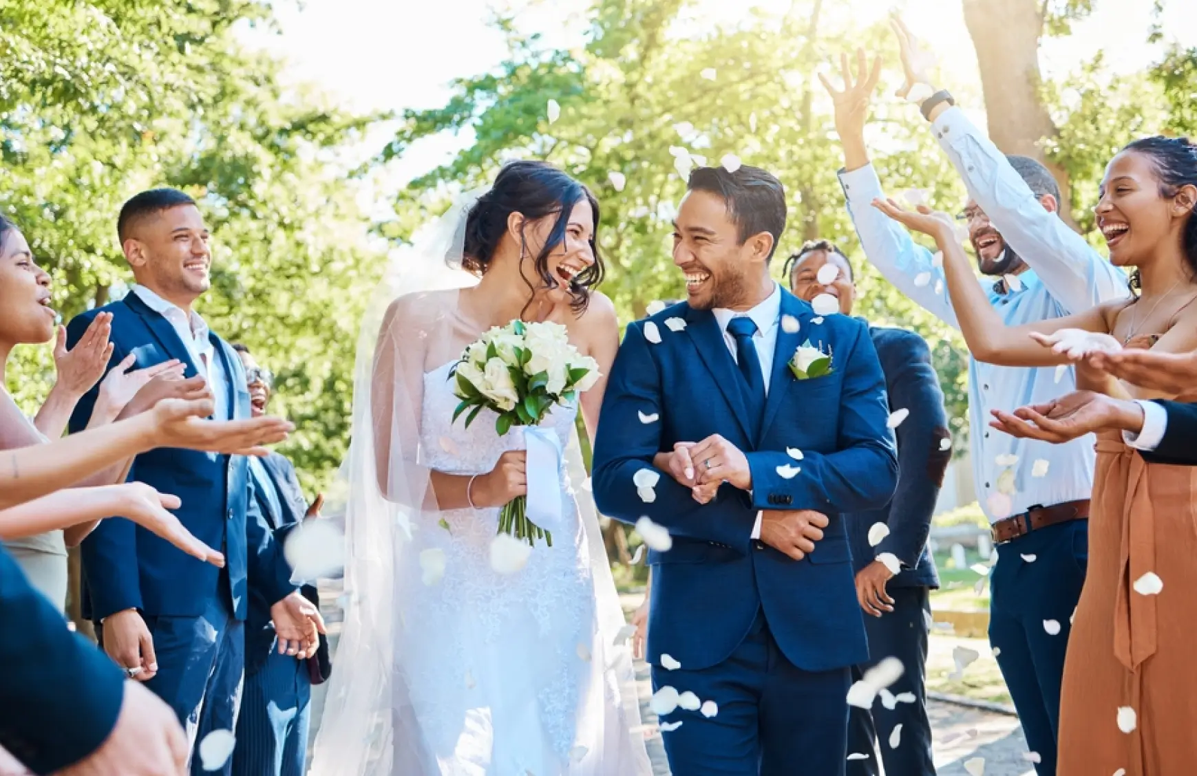 A newlywed couple walks down the aisle outdoors while guests cheer and shower them with flower petals.