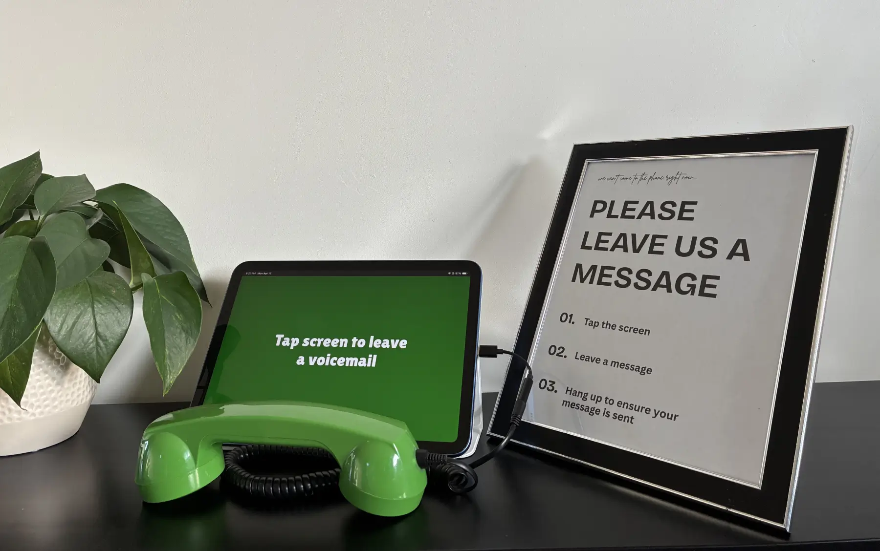 A GuestCam audio guestbook PhoneBooth setup with a tablet in recording mode, a green retro phone handset, and a framed 'Please Leave Us a Message' instruction sign.