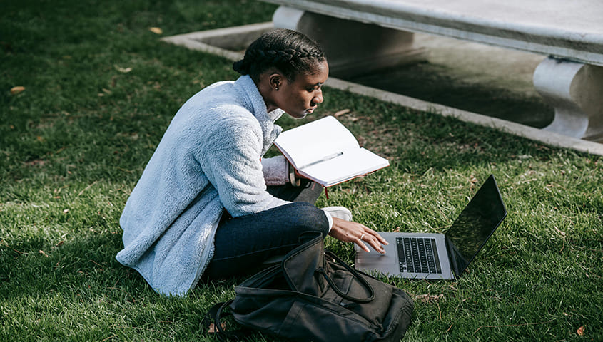 Woman sitting on grass researching Printful alternatives on a laptop
