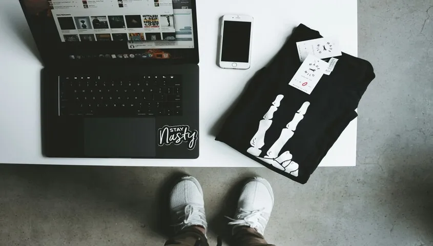A creative studio desk with a laptop and a custom-printed black tee, representing the workflow of t shirt printing and managing a fashion brand online.