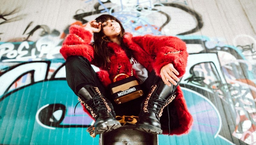 A woman sits confidently against a graffiti-covered wall wearing a bold red faux fur coat, black combat boots with leopard print detailing, black pants, and a Moschino handbag, looking directly into the camera.