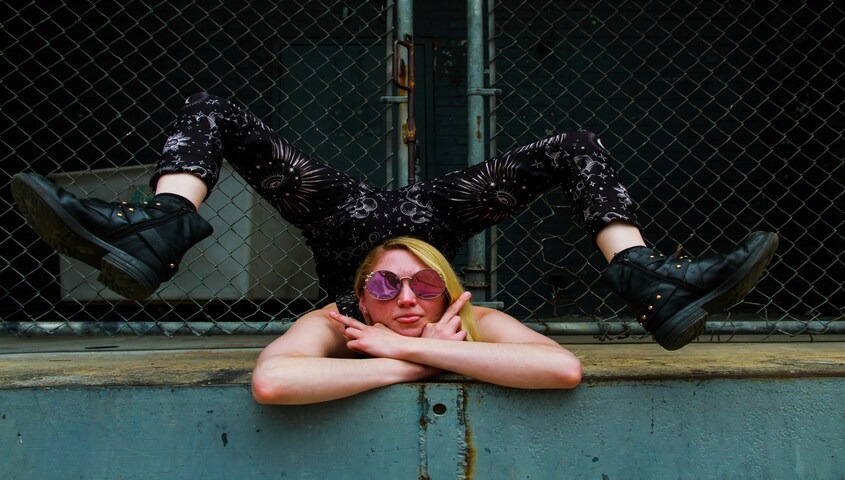A blonde woman with pink tinted round sunglasses rests her chin on crossed arms over a weathered teal ledge, legs kicked up behind her against a chain-link fence, wearing black platform boots and dark floral-patterned pants.