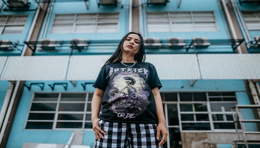 Woman in grunge fashion outfit — oversized band tee and checkered pants — standing in front of a blue urban building, embodying the grunge aesthetic