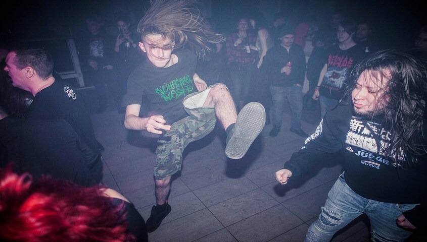 A young concertgoer with long hair mid-mosh at a dimly lit underground show, wearing a black band t-shirt, camo shorts, and black sneakers, surrounded by other attendees in dark clothing.