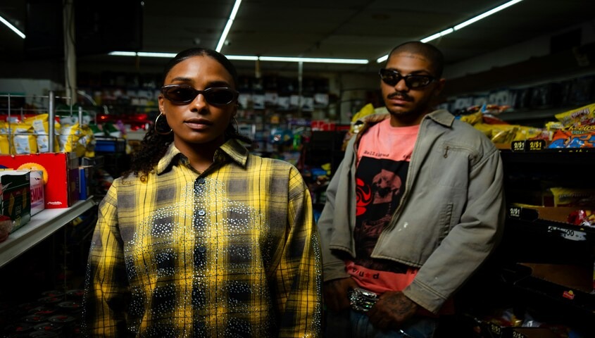 Two people wearing fashion-forward streetwear in a convenience store setting, representing the aesthetic of an independent clothing brand built on custom apparel blanks