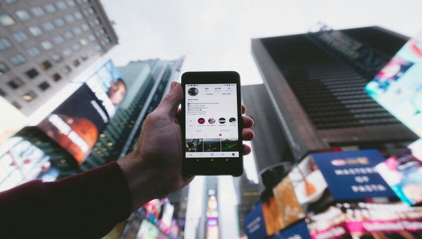 Hand holding a smartphone displaying an Instagram profile against a backdrop of city skyscrapers, illustrating social media management as an online business model