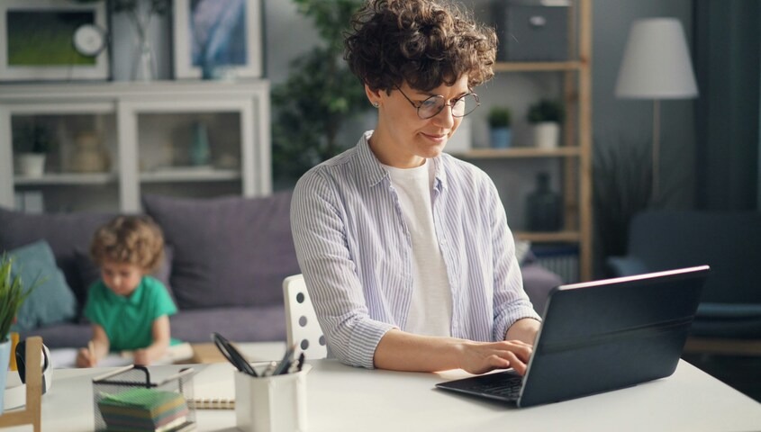 Stay at home mom blogging on her laptop while her child draws in the background, a long-term way to make money as a stay at home mom from home