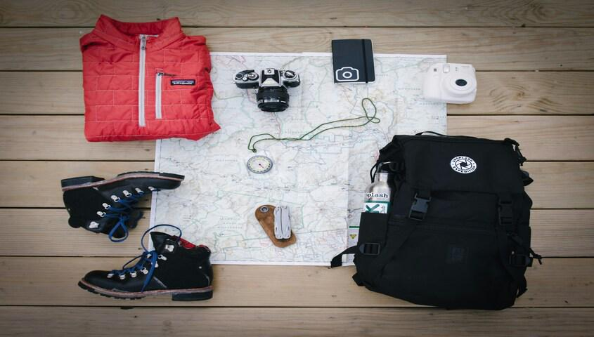 Flat lay of gorpcore fashion essentials including a quilted jacket, hiking boots, a compass, and a backpack.