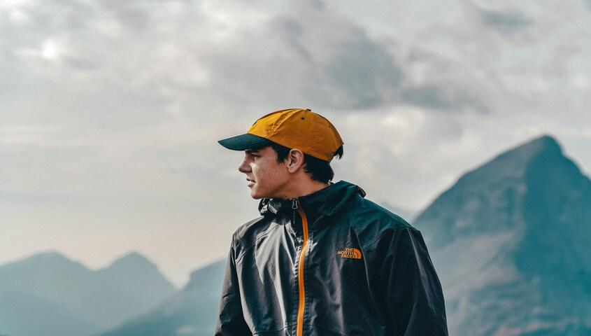 Man in a yellow cap and technical shell jacket in front of misty mountains, gorpcore fashion worn as an everyday default