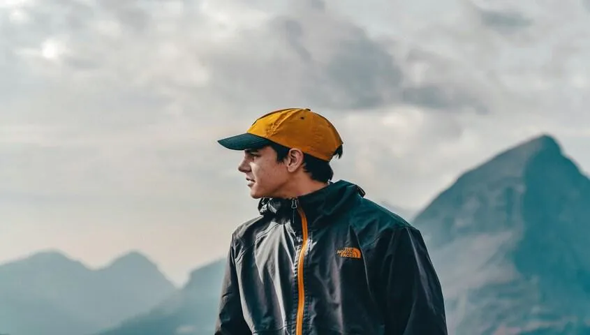 Man in a yellow cap and technical shell jacket in front of misty mountains, gorpcore fashion worn as an everyday default