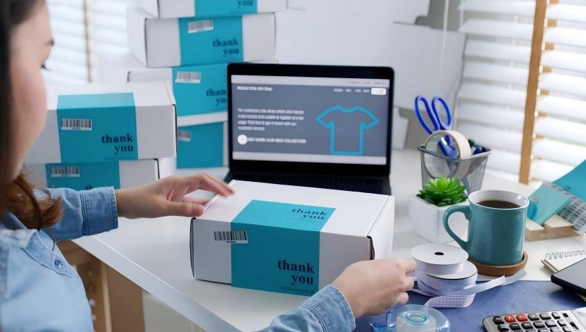 A female seller packing a “thank you” labeled shipping box at a desk beside a laptop displaying a T-shirt design.