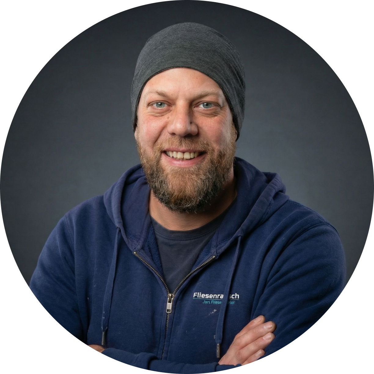 Lächelnder Mann mit Bart und grauer Mütze in einem dunkelblauen Kapuzenpullover vor dunklem Hintergrund.