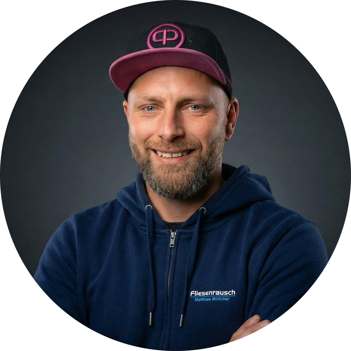 Mann mit Bart und blauen Augen trägt eine blaue Kapuzenjacke mit der Aufschrift Fliesenrausch Matthias Böttcher und eine schwarze Kappe mit pinkem Schirm und Logo.