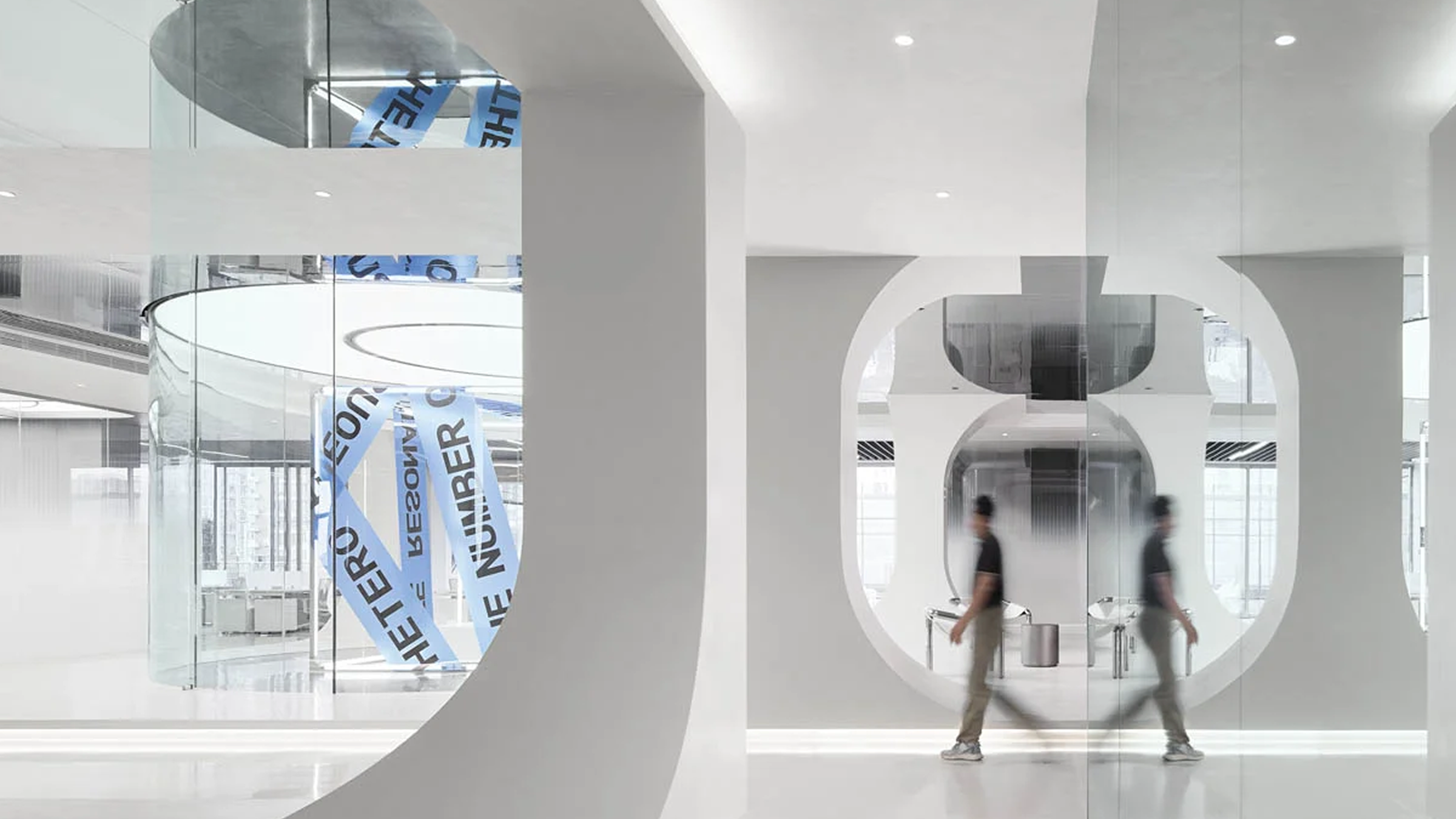 Modern office interior with curved white walls, glass partitions, and a blurred person walking past a reflective surface.