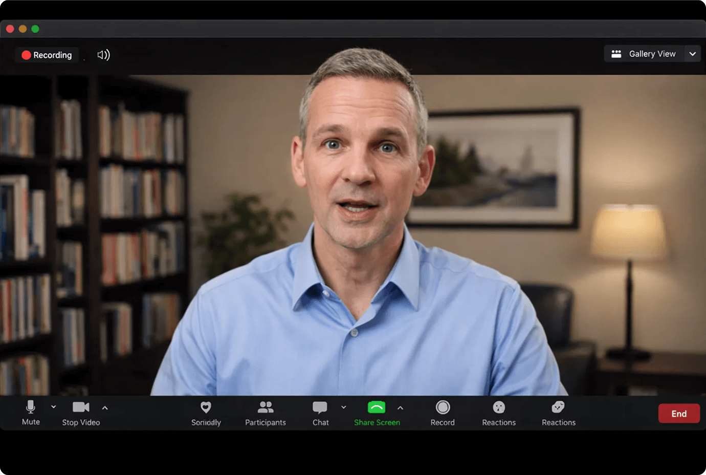 Middle-aged man with short gray hair in a blue shirt speaking during a video call with a bookshelf and a lamp in the background.