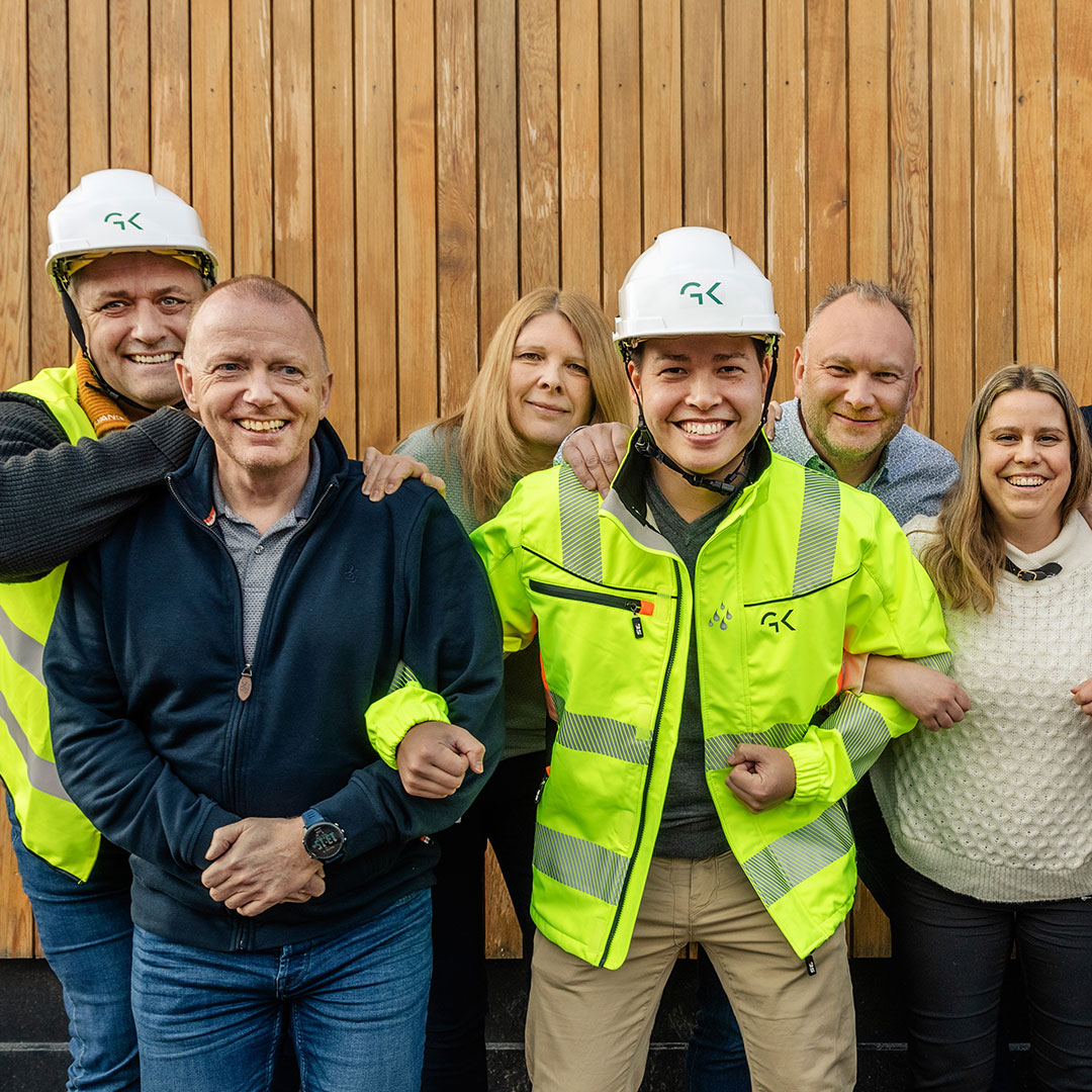 Sex personer, inklusive två i varselvästar och hjälmar med GK-logotyp, ler och står framför en träpanelvägg.