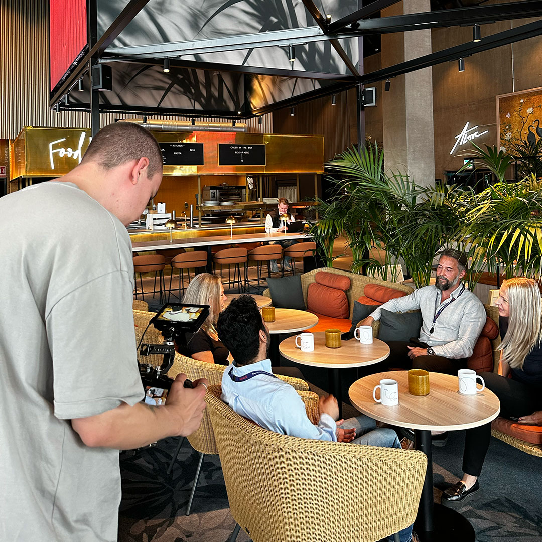 En person hanterar en kamera medan fyra personer sitter och samtalar runt bord i en modern kafémiljö med gröna växter och guldaccents.