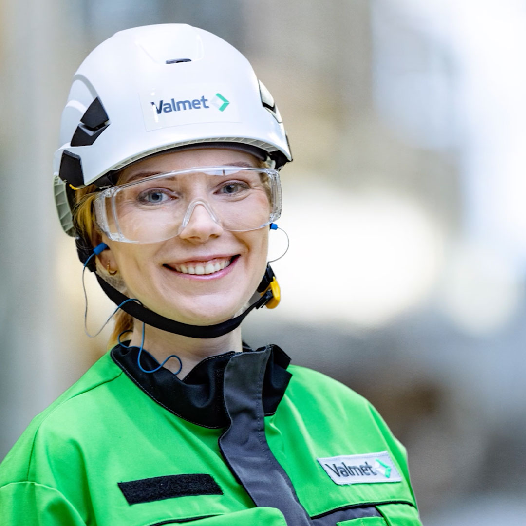 Leende kvinna i grön skyddsklädsel och vit hjälm med Valmet-logotyp på en arbetsplats.