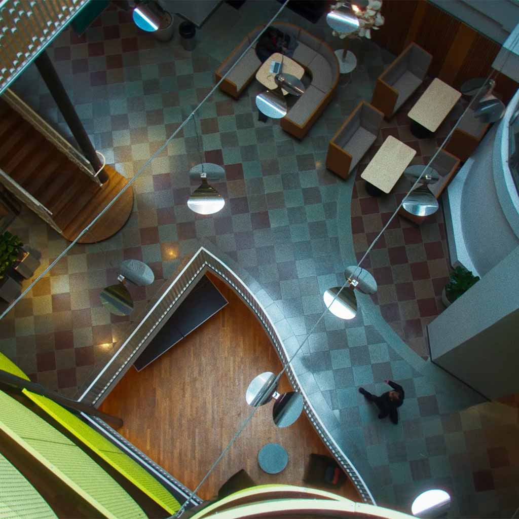 Vy uppifrån över en modern inomhuslobby med mönstrat golv, hängande lampor, soffor, bord och en person som går.