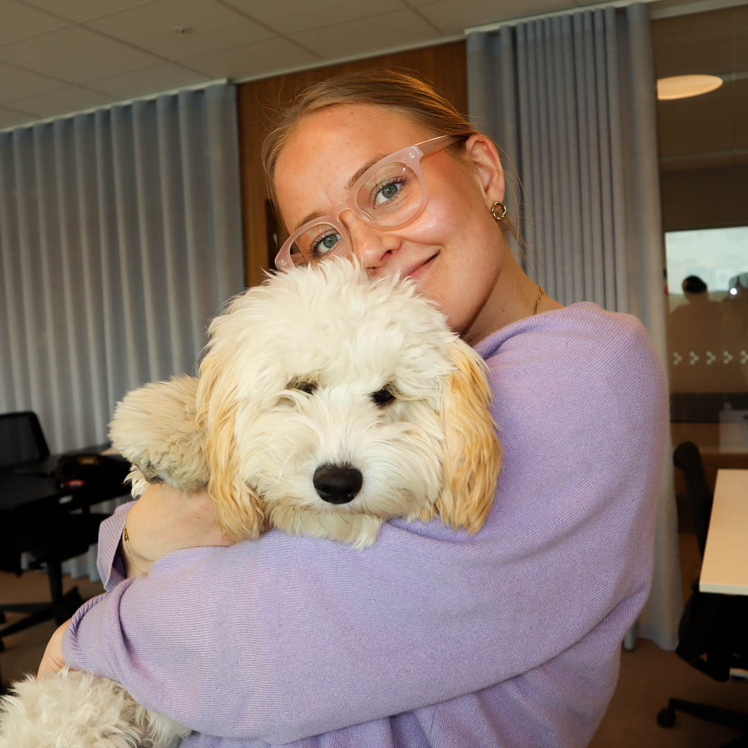 Kvinna med glasögon i lila tröja håller om en fluffig vit hund inomhus.