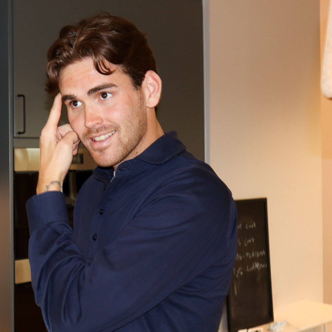 Man med mörkt hår och mörkblå skjorta pekar mot sitt huvud och ler.