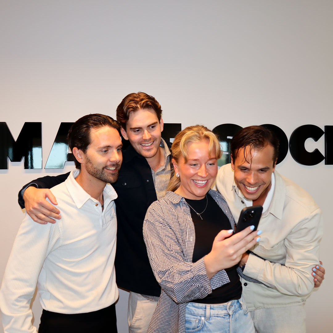 Fyra unga vuxna som ler och tar en gruppselfie inomhus framför en vägg med svart text.