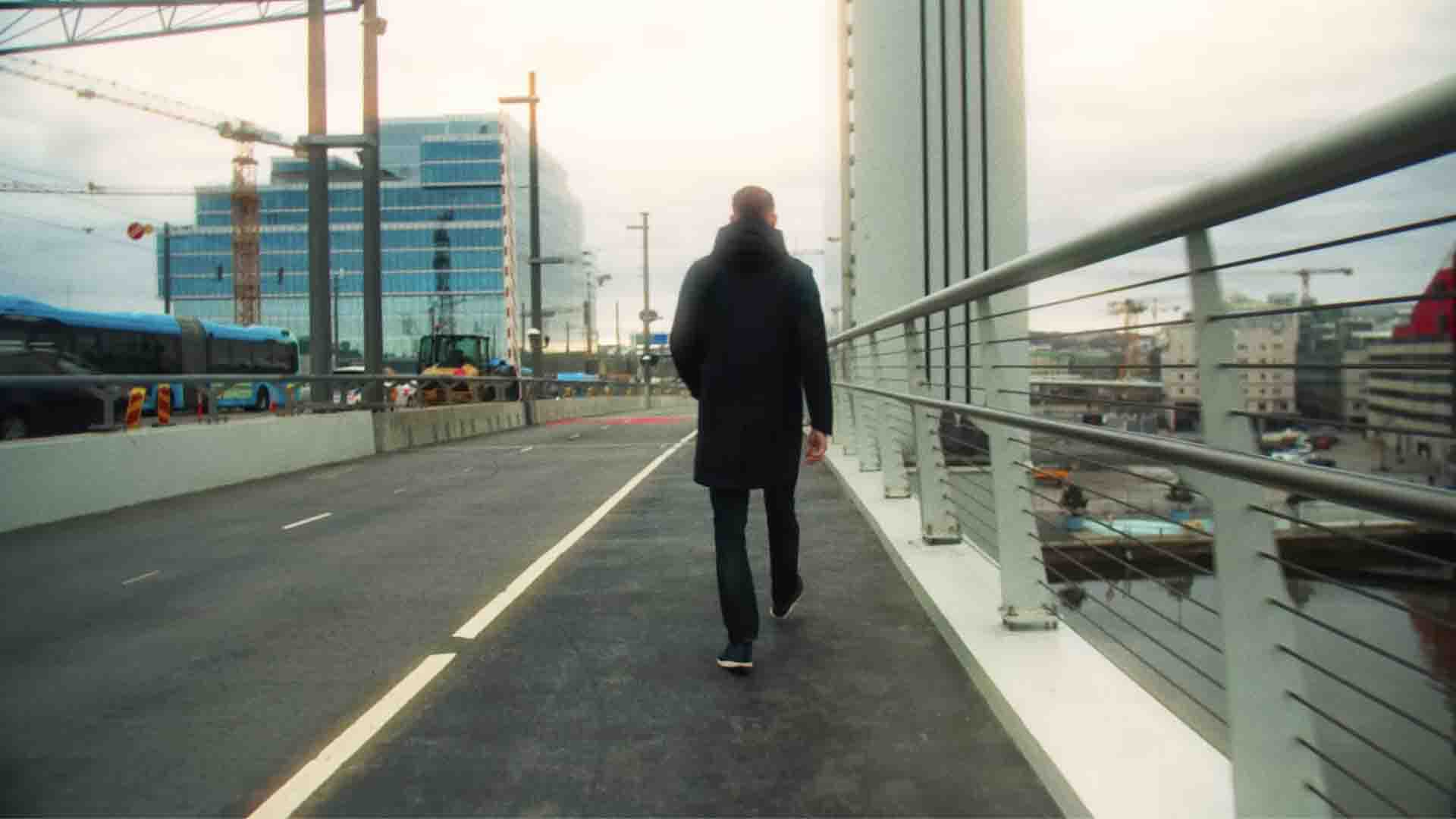 En man i svart rock går på en modern stadsgång med byggnader och byggkranar i bakgrunden.