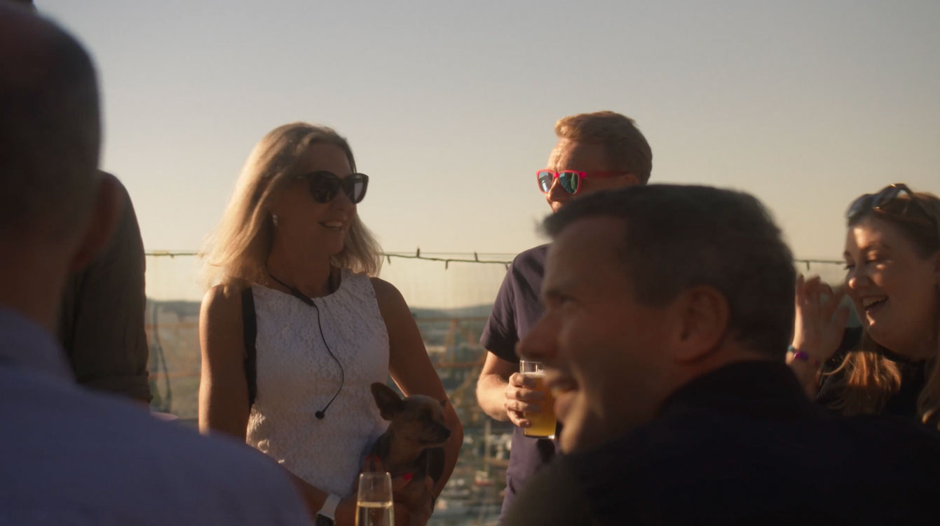 En grupp människor utomhus i solnedgång, en kvinna håller en liten hund medan de pratar och skrattar.