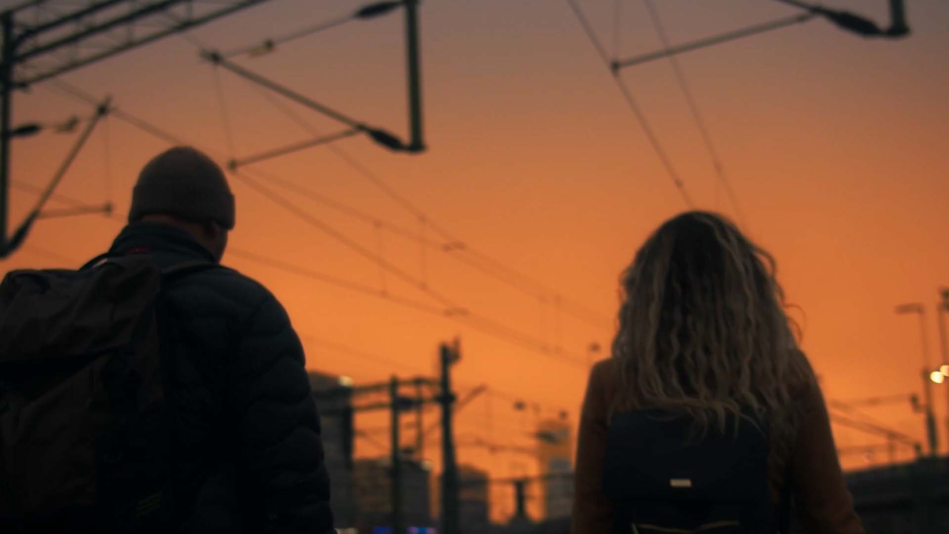 Två personer med ryggsäckar står vid en tågstation under en orange solnedgång.