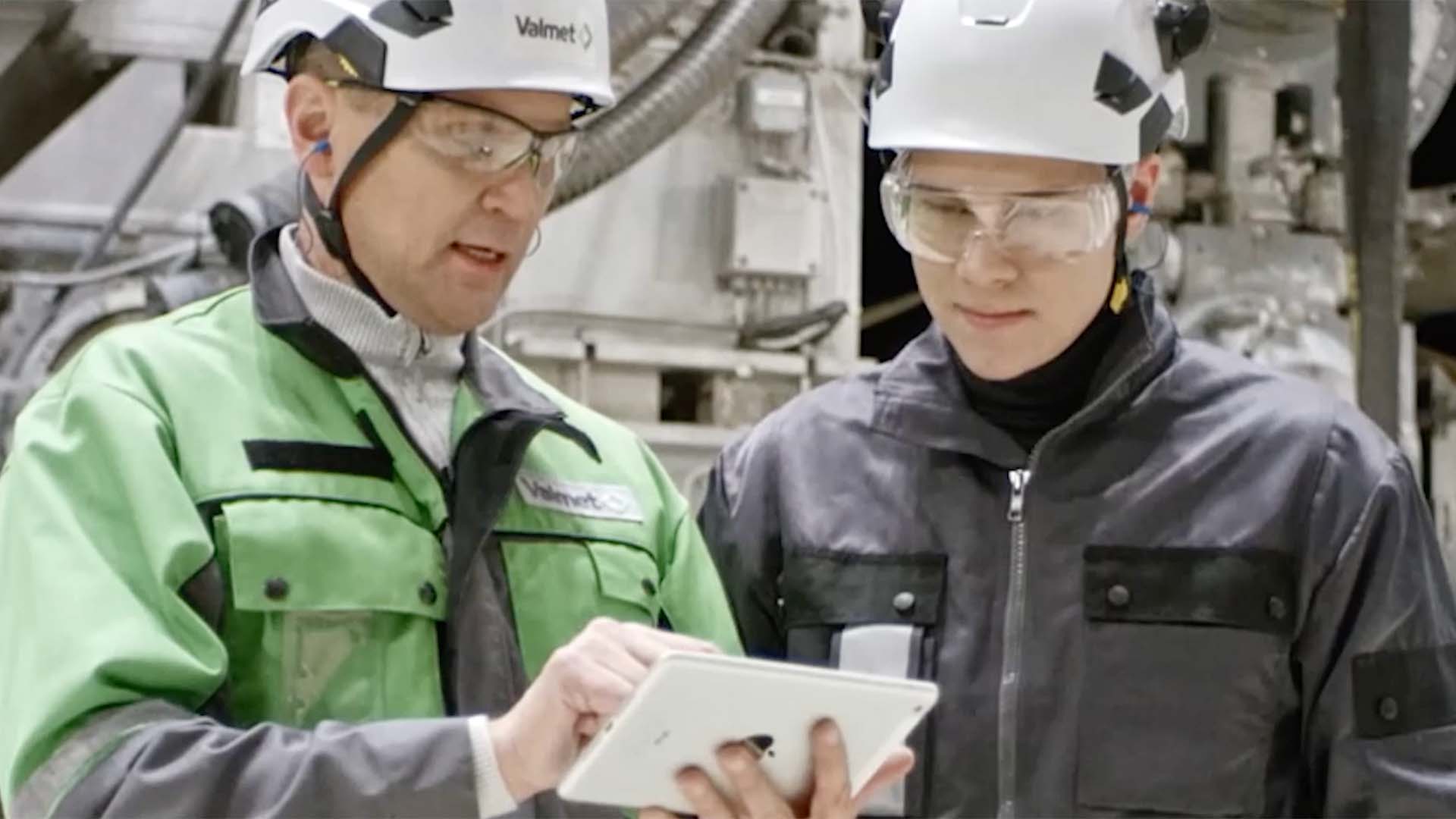 Två fabriksarbetare i skyddshjälmar och glasögon granskar en surfplatta i en industriell miljö.