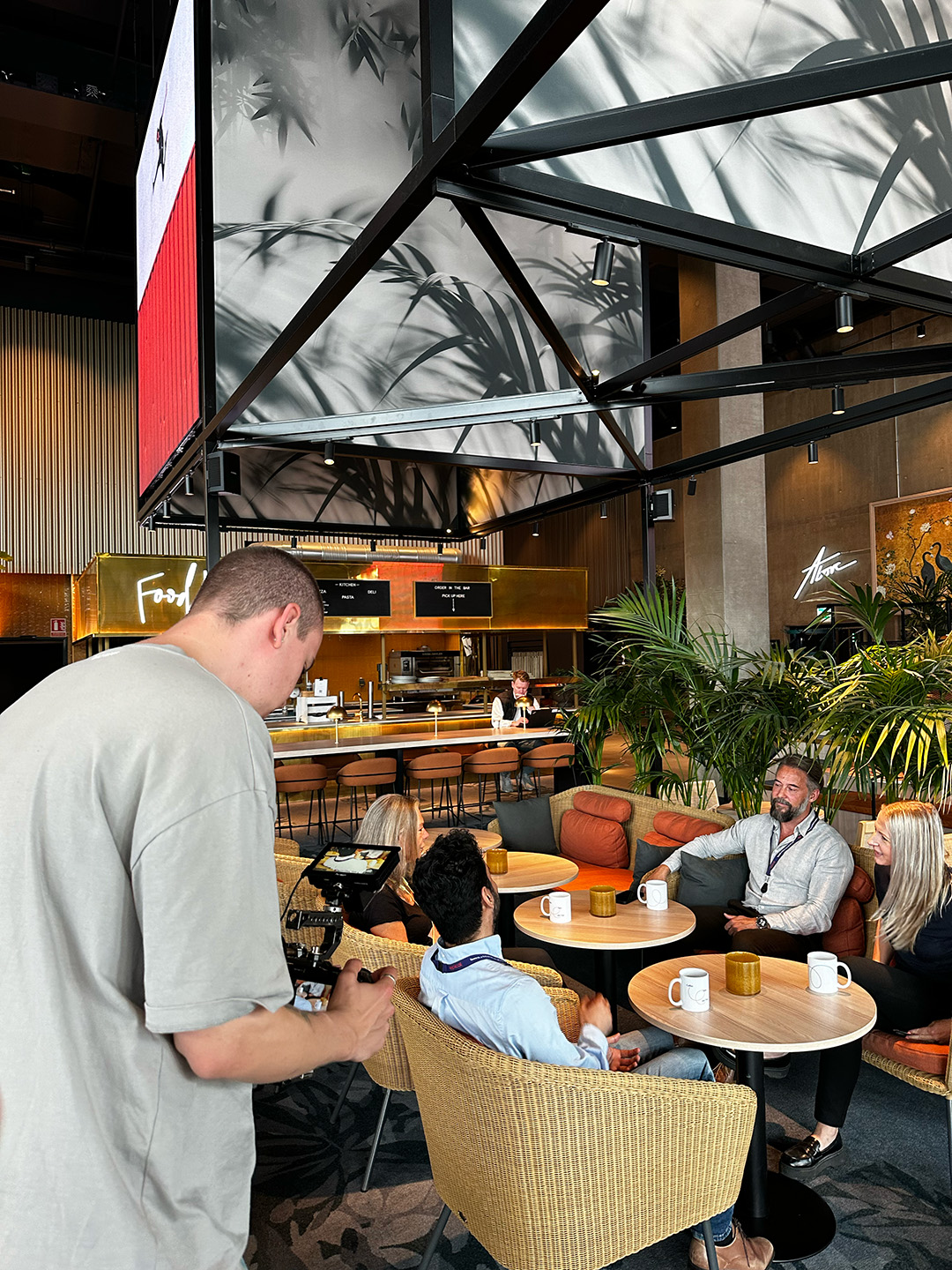 Fyra personer sitter och samtalar i en lounge med rottingstolar och runda bord, medan en person med kamera filmar dem i en modern kafé- eller kontorsmiljö.