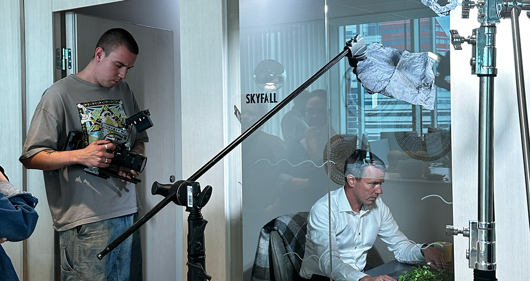 En man står med en kamera rigg medan en annan man sitter och arbetar vid en dator i ett glasrums-kontor med skylten Skyfall.