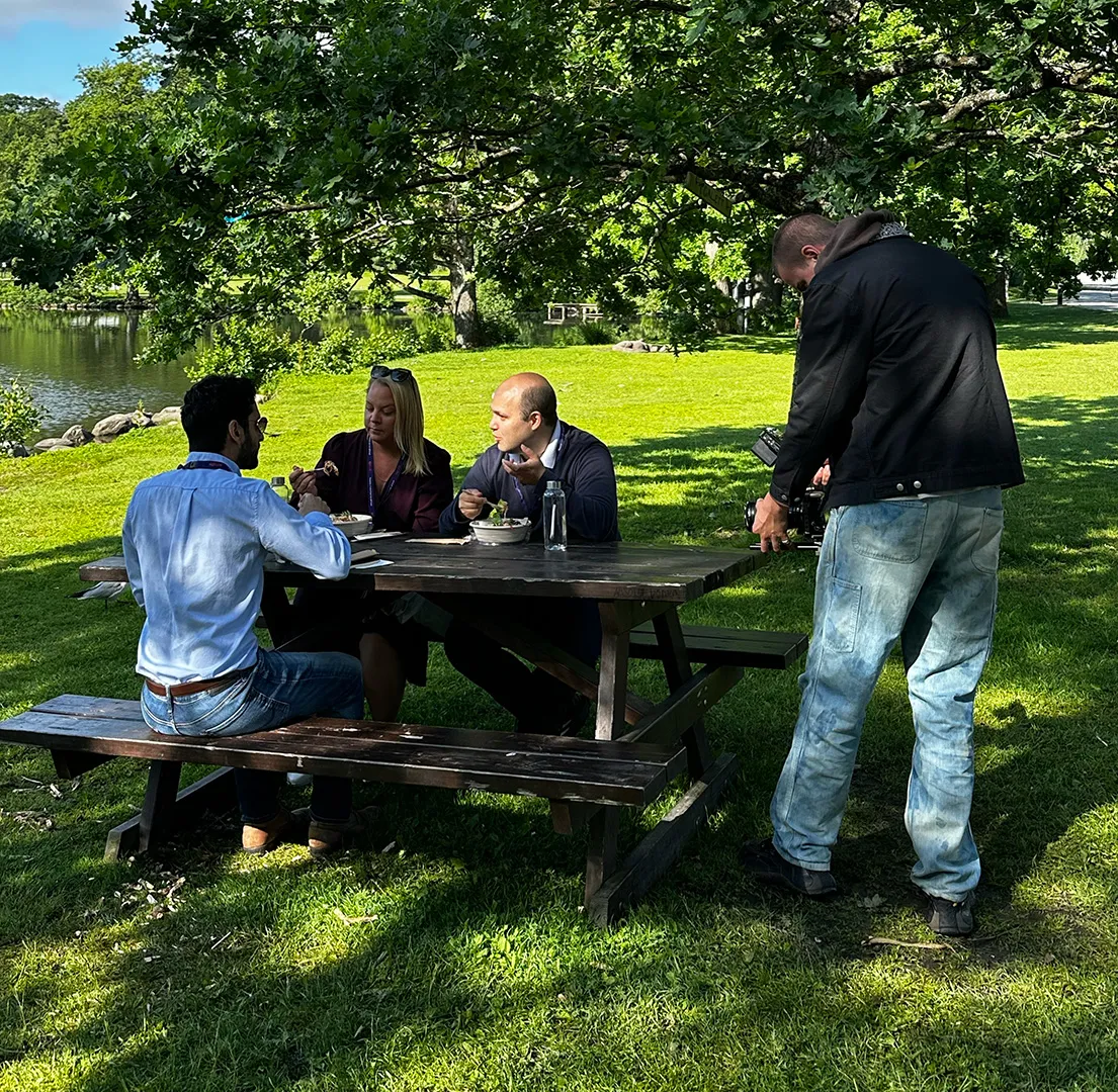 Tre personer sitter vid ett träbord i en park och äter mat medan en person filmar dem med en kamera.