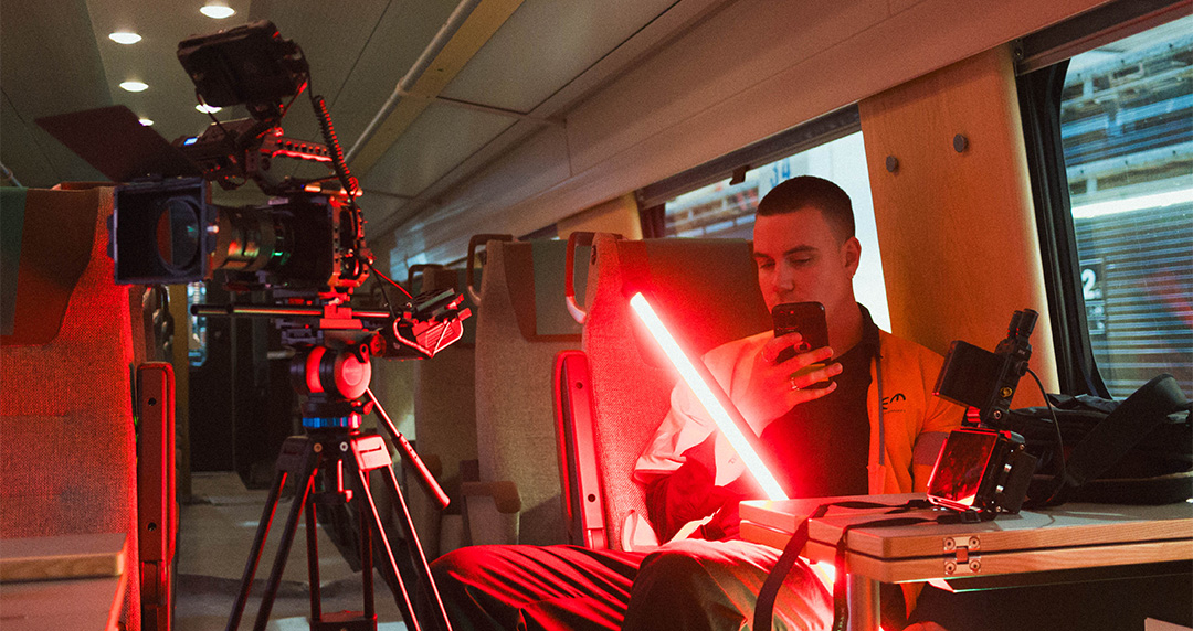 Person sitter på ett tåg med filmutrustning runt sig och håller i en röd ljusstav medan de tittar på sin mobiltelefon.