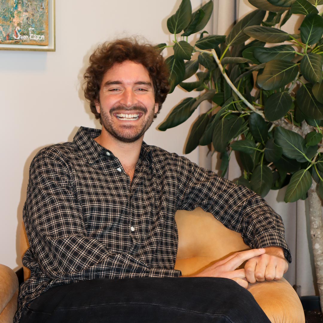 Leende man med lockigt hår och skägg i rutig skjorta sitter avslappnat i en fåtölj med en stor grön växt i bakgrunden.