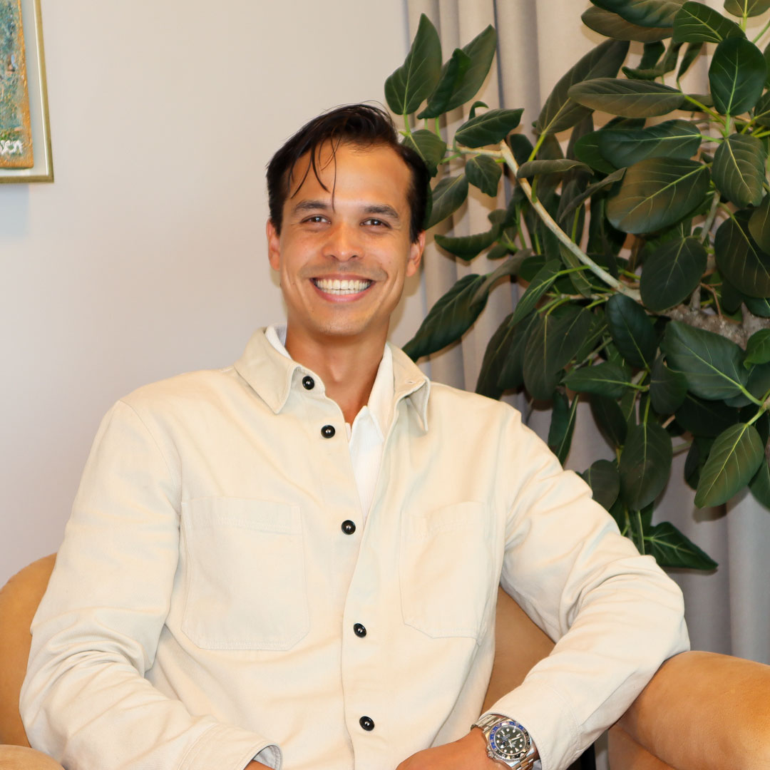 Leende man i ljus jacka sitter avslappnat i fåtölj med grön växt i bakgrunden.