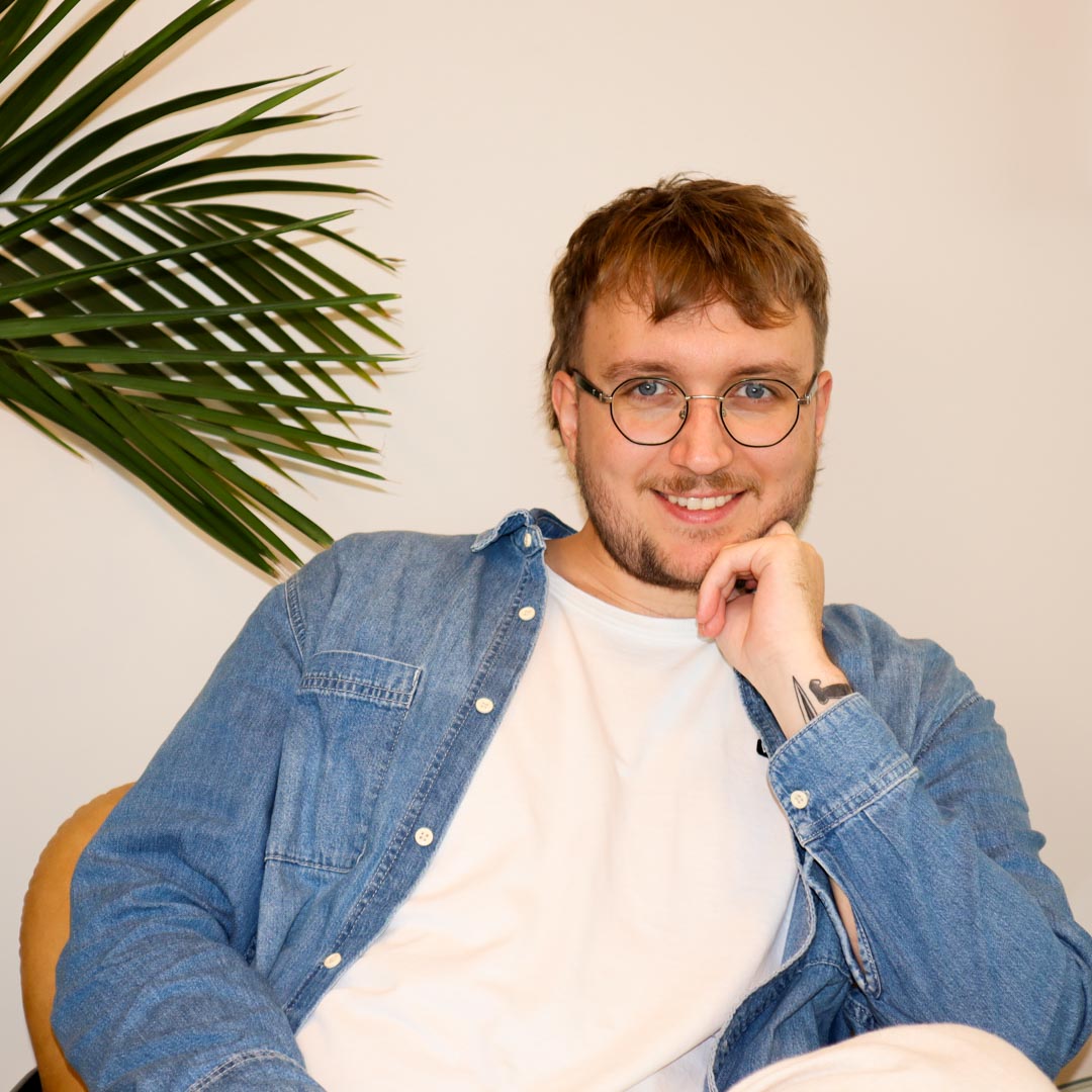 Man med glasögon och skägg, iklädd vit t-shirt och jeansjacka, sitter avslappnat med handen nära hakan framför ljus bakgrund och grönt växtblad.