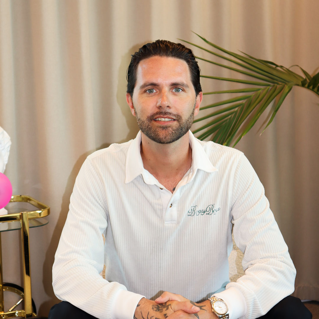 Man med mörkt hår och skägg, iklädd vit pikétröja och guldklocka, sitter framför beige gardin och en grön växt.