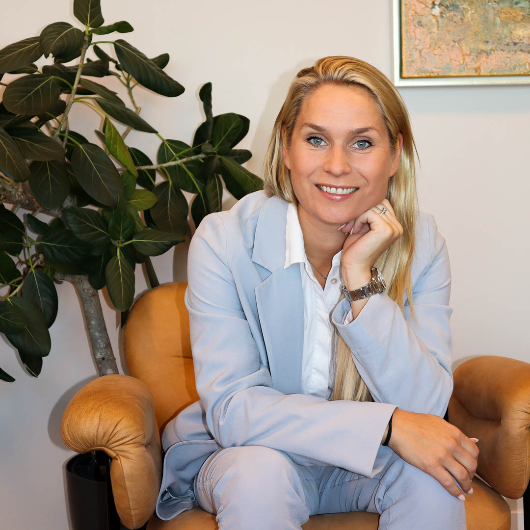 Kvinna med långt blont hår i ljus kostym som sitter i en beige stol med en grön växt i bakgrunden.