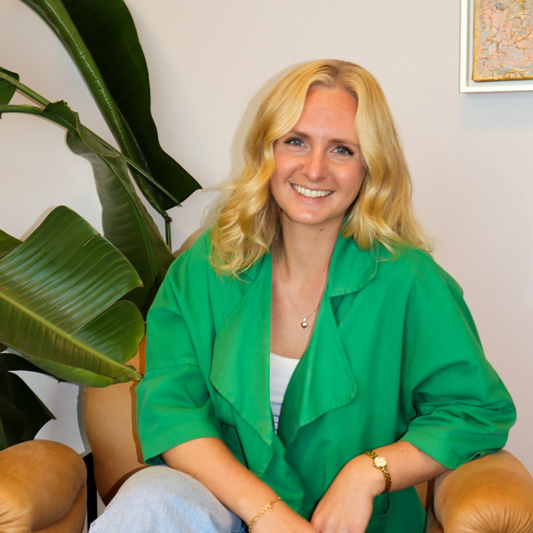 Leende kvinna med blont hår i grön kappa sitter i läderfåtölj bredvid stora gröna växtblad.