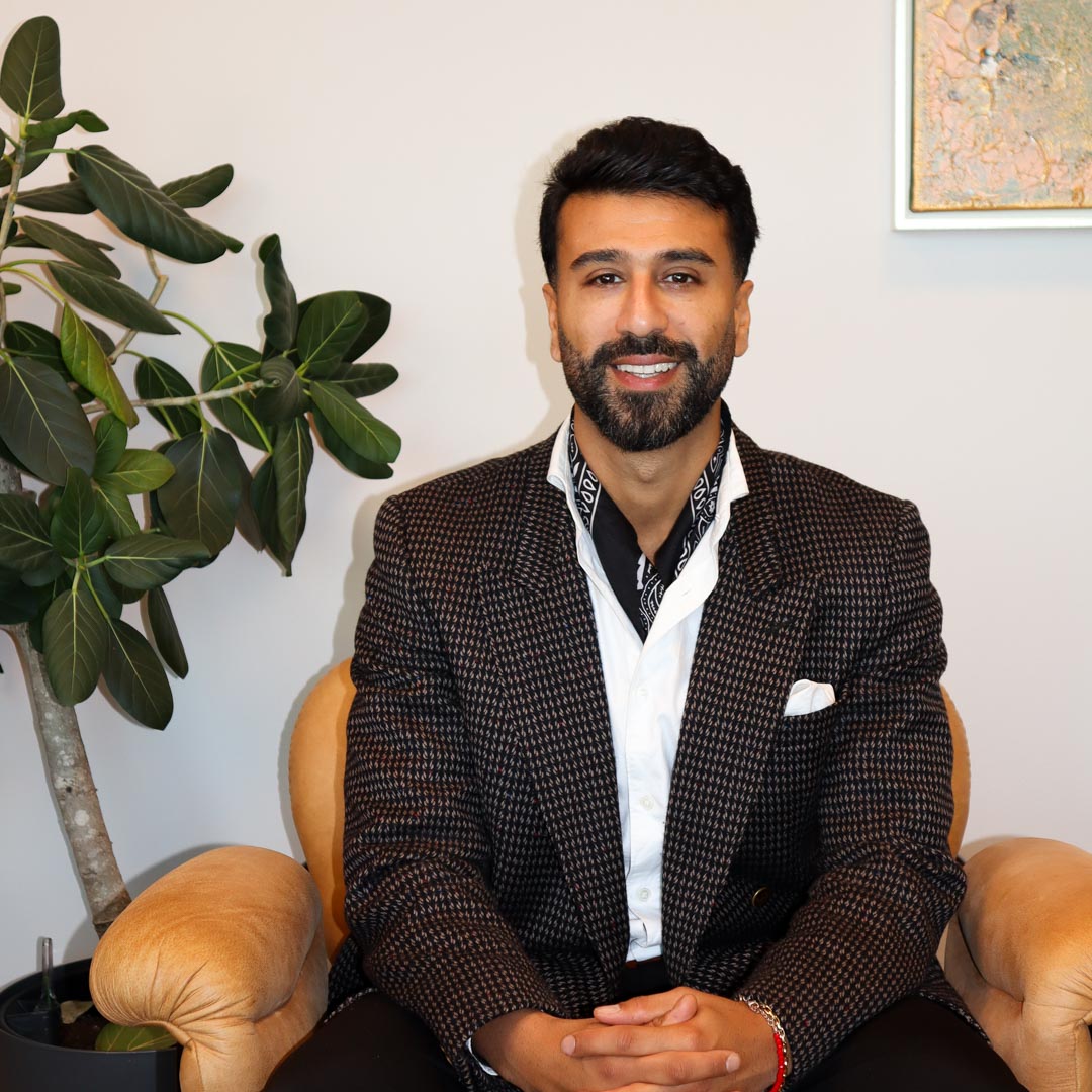 Man med skägg och svartvitt halsduk sitter på en beige fåtölj bredvid en stor grönt bladig växt.