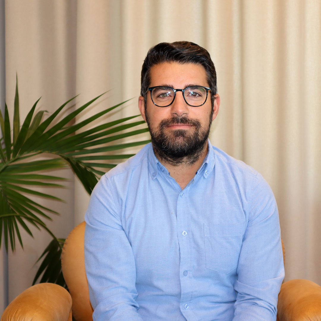 Man med mörkt hår, glasögon och skägg i ljusblå skjorta sitter i en beige fåtölj med gröna palmblad bakom.