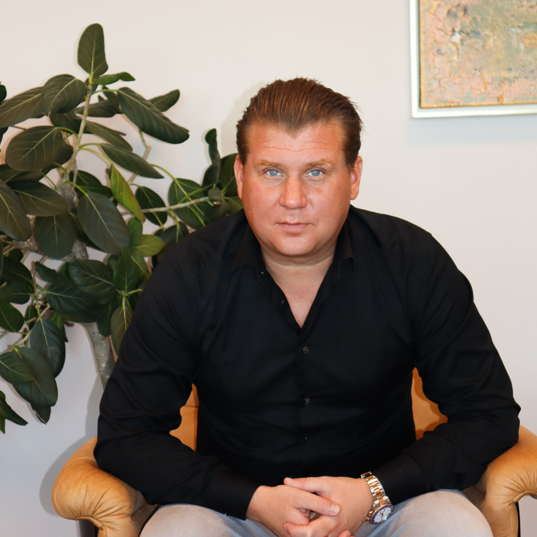 Man med blå ögon sitter i en fåtölj framför en växt och en målning på väggen.