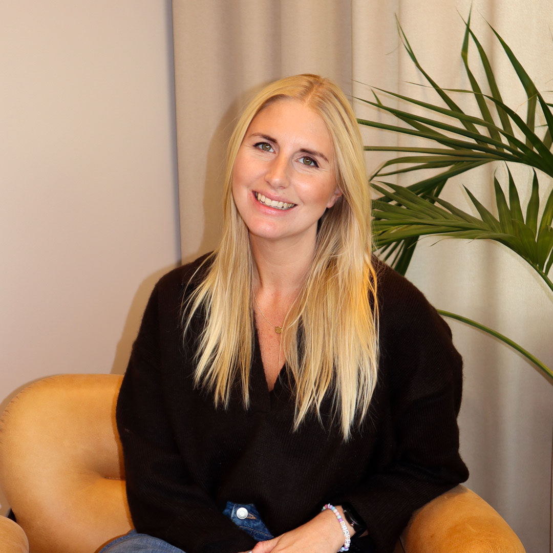 Leende kvinna med långt blont hår, svart tröja och blå jeans sitter i en beige fåtölj med gröna växtblad i bakgrunden.